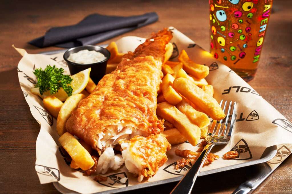 photograph of Traditional fish and chips, on a metal tray with lemon wedge, tartare sauce, parsley garnish and a pint of neck oil ale in the background