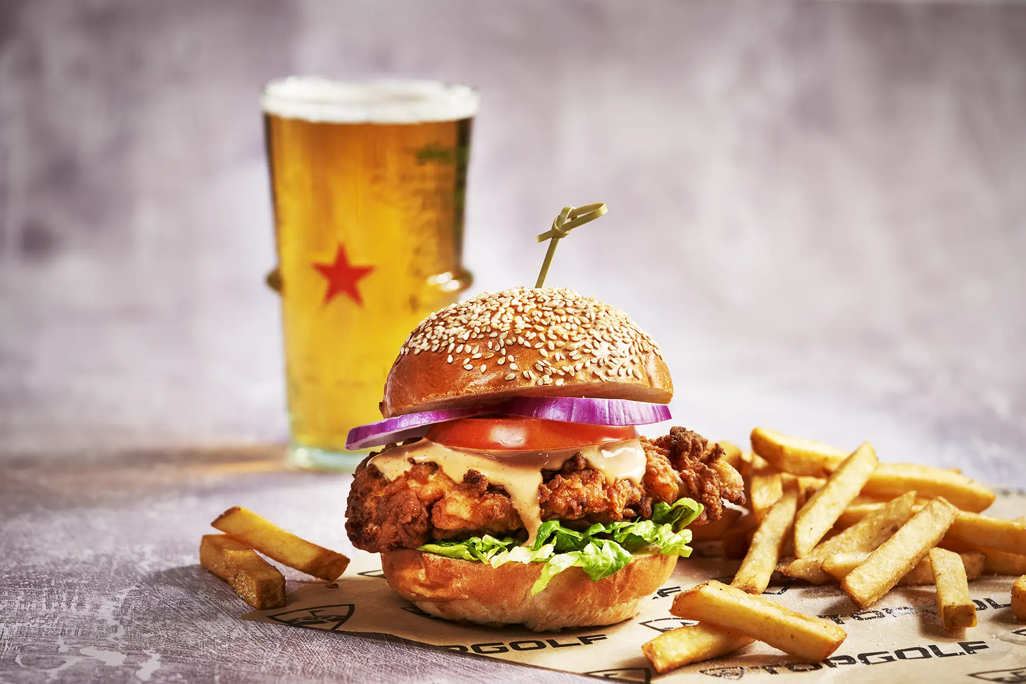 Chunky chicken burger with mayo, tomato, red onion and lettuce in a toasted sesame seeded bun with fries and an ice cold pint of beer in the background.